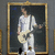 A framed portrait of a person in white clothing holding a guitar, set against a dark background with blue accents.