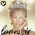 Pixelated image of a person with light brown hair wearing a tiara, smiling while holding a microphone near their mouth.