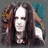 A static image of a person with long dark hair and white face paint, wearing a black outfit against a teal background.