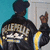 A person in a black jacket with yellow stripes and 'FREEDOM' text on the back, standing against a teal wall and white door, making a hand gesture.
