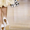 A cropped view of a person's lower body wearing a light yellow skirt and white high-heeled shoes, holding a black-and-white checkered handbag.