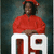A person in a red graduation gown and cap sits holding large white '09' numerals against a dark blue background.