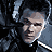 Pixelated static image of a person with dark hair and light skin, wearing a black top against a dark background.