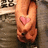 A pixelated close-up of a light-skinned hand making a peace sign with the index and middle fingers, holding a small pink heart-shaped object against a blurred background.