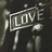 A monochrome icon with a sign reading 'LOVE' in block letters, surrounded by blurred shapes resembling hands or fingers against a dark background.
