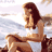 Pixelated static image of a woman in a white tank top sitting on rocky terrain with water and sky background.