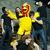A pixelated image shows a person in a yellow mascot costume with red accents, arms raised among a group of people. Text reads "MAYDAY PARADE!!" and "love".