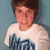 Pixelated image of a person with brown hair wearing a white shirt with dark blue text against a light blue background.