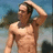 A shirtless man with short dark hair stands outdoors, hands behind his head, against a blurred background of blue sky and trees.