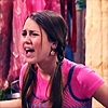 A person with long brown hair in a braid wears a pink shirt with blue trim, standing against a pink and red patterned backdrop.