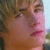 Pixelated close-up of a person's face with light brown hair, pink lips, and visible eyes against an outdoor background.