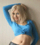 A person with short blonde hair wearing a blue long-sleeve crop top and dark shorts poses with hands behind head against a light wall.