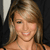 A pixelated close-up of a smiling person with light brown hair, wearing a thin necklace and dark straps, against a dark background with a light vertical element.