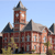 Pixelated image of a red-brick building with a tall tower, dark roof, and light-colored windows under a clear blue sky.