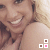 A pixelated close-up of a smiling person with light brown hair, wearing a strapless top and holding their shoulder.