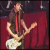 A person with long brown hair and red necklaces plays a white electric guitar against a dark background with red curtains.