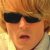 Pixelated close-up of a person with light brown hair wearing dark sunglasses and a light-colored top against a warm-toned background.