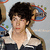 Pixelated static image of a person with dark curly hair wearing white and black clothing, red lips, and a blue-and-red circular object behind the head.