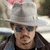 A person wearing a light gray wide-brimmed hat and dark sunglasses, with a mustache and goatee, in front of blurred background elements.