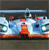 Pixelated static image showing a stylized vehicle with blue and white body, orange accents, and dark wheels on a gray background.