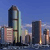 Pixelated cityscape with tall buildings against a blue sky, featuring a prominent central tower with reflective glass and a dark top section.