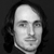Static grayscale image of a person's face with long dark hair, neutral expression, and visible neck against a dark background.