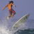 Pixelated image of a person surfing on a wave with a light blue sky background, wearing an orange shirt and red shorts.