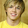 A person with light brown hair wearing a yellow shirt against a blurred background.