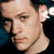Pixelated close-up of a person's face with light skin tone, dark hair, and a thin chain necklace against a dark background.