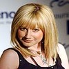 A static image of a person with shoulder-length blonde hair and bangs, wearing a black sleeveless top and a silver necklace with a pendant, against a blurred background with partial text.