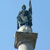 Pixelated statue of a figure with raised arm on a pedestal against light blue background