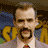 A pixelated image of a man with short brown hair and a beard, wearing a light gray suit jacket over a white shirt and red tie.