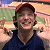 Pixelated image of a person wearing a dark blue shirt and red cap, standing against a blurred background with green and orange tones.