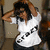 A person in a white shirt with black text stands against a blurred background, hands raised to their head across four