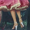 A close-up of a person's lower legs and feet wearing a pink skirt with gold trim and white high-heeled shoes with ankle straps.