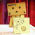 A pixelated Danbo character holds a yellow card with a flower and coin design against a red curtain background.