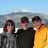 Three people stand side-by-side against a blurred background of water and sky, wearing dark and yellow clothing with pixelated facial features.