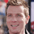 Pixelated close-up of a person with light brown hair, fair skin, and a slight smile, wearing a dark jacket with a white collar.