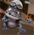 Pixelated silver robot with large eyes, rounded body, and extended arm holding a small orange object against a brown background.