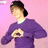 Pixelated figure in purple sweater with white collar, dark hair tied back, standing against pink background holding circular object.