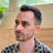 Pixelated static image of a person with short dark hair, light skin, and stubble wearing a black-and-white patterned shirt against a blurred indoor background.