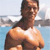 Pixelated image of a shirtless person with dark hair standing near water, background includes distant structures and sky.