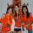 Three people in orange shirts and pumpkin hats pose together against a light background, with text on an adjacent orange