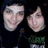 Two people in dark hooded clothing with green and white text on the chest, smiling and neutral expressions in a