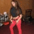 A person in a black top and red pants stands in a room with a brown wall, holding a guitar-like object, with a chair and a dog visible in the background.