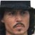 Static pixelated image of a person wearing a dark hat with long dark hair visible on the sides, set against a dark background.
