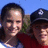 Two people pose outdoors under a blue sky, one wearing a dark cap with white emblem and red shirt.