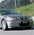Pixelated image of a silver sedan with dark tinted windows parked on a paved surface, green grassy hill in background, blue water body visible behind vehicle.