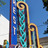 Pixelated static image of a vertical blue sign with white text and a golden scroll design on a blue sky background.