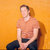 A pixelated figure in a light-colored shirt and dark pants sits against a solid orange background with a shadow to the right.